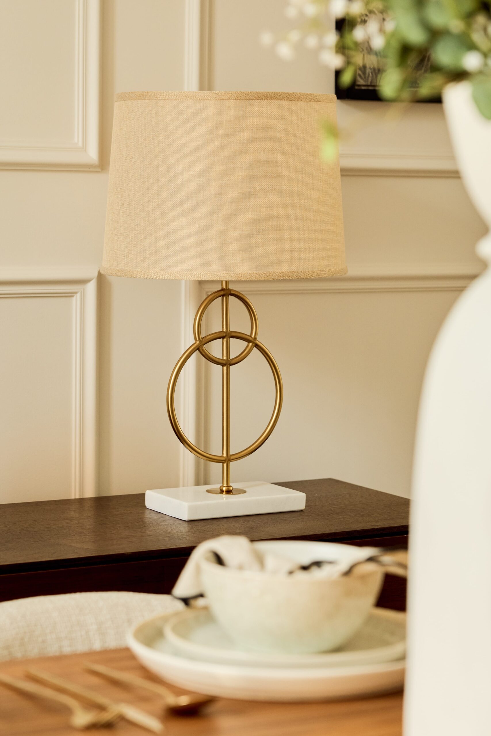 close up of gold sculptural lamp with beige lampshade against neutral background atop wooden sideboard